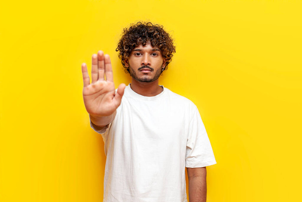 jeune homme indien en t-shirt blanc interdit et montre geste d'arrêt sur fond jaune isolé, bouclé gars montre la paume avertit et le refus - Photo, image