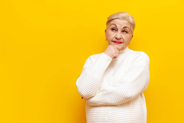 vieille grand-mère coûteuse dans un pull blanc pense et plans sur un fond jaune isolé, femme âgée pensionnée perplexe imagine et regarde loin - Photo, image