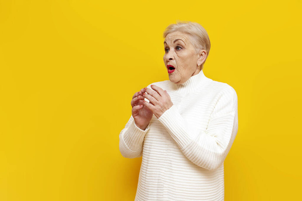 anciana asustada abuela en un suéter blanco se sorprende y se preocupa en un fondo aislado amarillo, sorprendido pensionista anciana mira hacia otro lado con asombro - Foto, imagen
