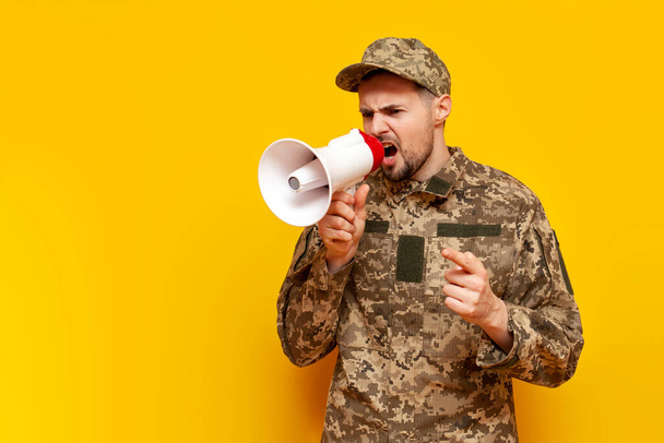 Soldado del ejército ucraniano en uniforme de camuflaje militar pixel anuncia información en un megáfono y grita sobre un fondo aislado amarillo, cadete militar ucraniano habla y advierte en un altavoz - Foto, imagen