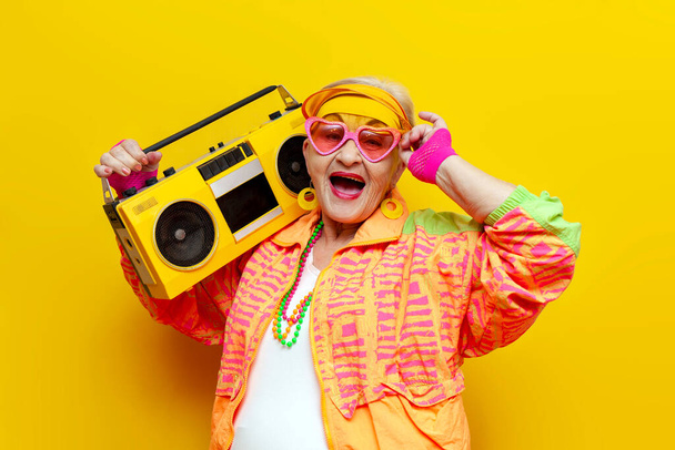 divertente nonna pazza in vestiti hipster ascoltare musica su registratore e cantare su sfondo giallo isolato, anziana donna fresca che balla alla alla festa e urla - Foto, immagini