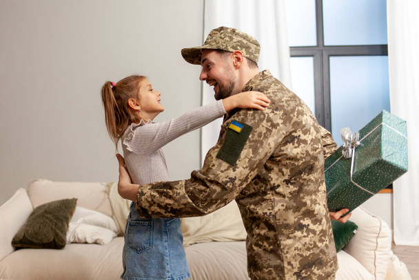 soldado feliz del ejército ucraniano en uniforme de camuflaje regresó a casa a su hija y la felicita en su cumpleaños, cadete militar da regalo a un niño para las vacaciones - Foto, Imagen