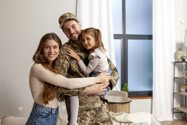 soldat heureux de l'armée ukrainienne en uniforme de camouflage retourné à la maison à sa famille, papa militaire rencontre et étreint sa femme et sa fille à la maison, concept de mobilisation en Ukraine - Photo, image