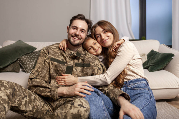 Ukrainian army soldier in camouflage uniform returned home to his family, military cadet sits on the sofa with his wife and daughter and smiles, child hugs veterans father - Photo, Image