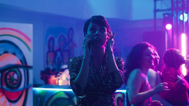 Woman answering phone call at party in the club, trying to have conversation with loud music on dance floor. Young adult using mobile phone remote chat at clubbing event, entertainment. - Photo, Image