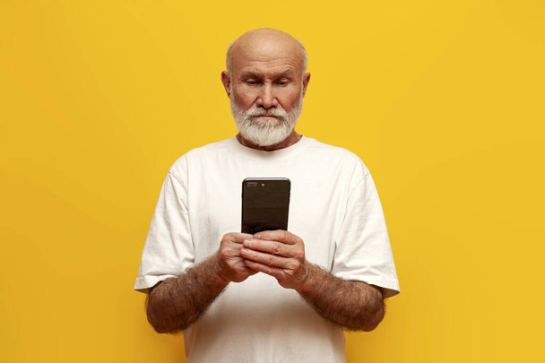 komoly öreg kopasz nagyapa fehér pólóban okostelefon sárga elszigetelt háttér, zavaros idős nyugdíjas gépelés online a telefonon - Fotó, kép