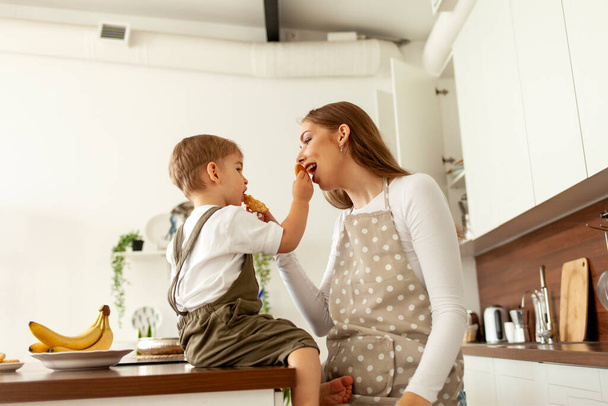 jeune mère en tablier donne des biscuits à son petit fils à manger dans la cuisine à la maison, garçon de 2 ans prend le petit déjeuner avec son parent, femme nourrit l'enfant - Photo, image
