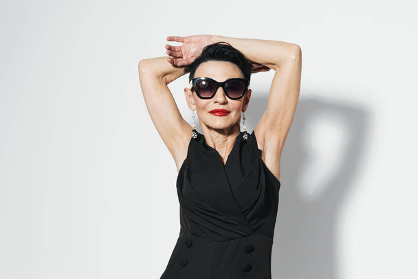 Femme mature élégante en robe noire et lunettes de soleil posant avec confiance avec les mains sur la tête pour la prise de vue de la caméra - Photo, image