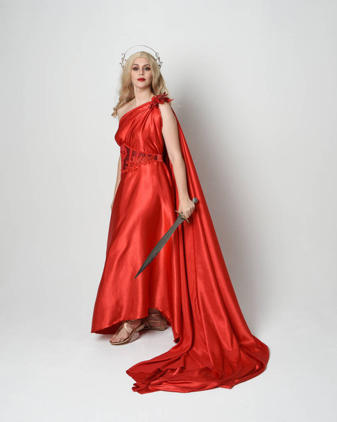 Täyspitkä muotokuva kaunis blondi malli pukeutunut antiikin mytologinen fantasia jumalatar virtaava punainen silkki tooga puku, kruunu. kävely aiheuttaa, miekka-ase, eristetty studio tausta - Valokuva, kuva