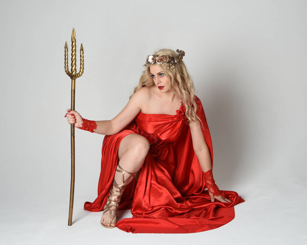 Portrait de pleine longueur d'un beau modèle blond habillé en ancienne déesse mythologique fantaisie en robe de toge de soie rouge fluide, couronne. pose à genoux, arme trident dorée, fond studio isolé - Photo, image