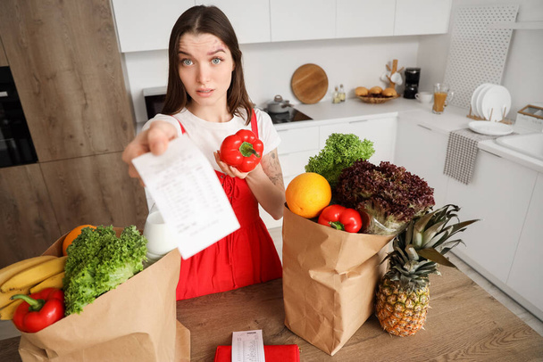 Σοκαρισμένη νεαρή γυναίκα με σακούλες φαγητού και απόδειξη καταστήματος στην κουζίνα. Έννοια της αύξησης τιμών - Φωτογραφία, εικόνα
