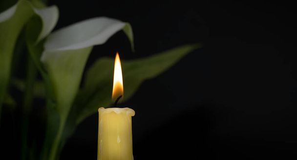 Una sola vela encendida toma protagonismo en el primer plano junto a ella lirios de calas blancas con hojas verdes que sugieren recuerdo, reflexión y esperanza - Foto, Imagen