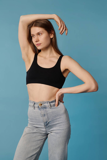 Vertical medium long studio portrait of young Caucasian woman wearing crop top and jeans posing for camera, blue background - Фото, изображение