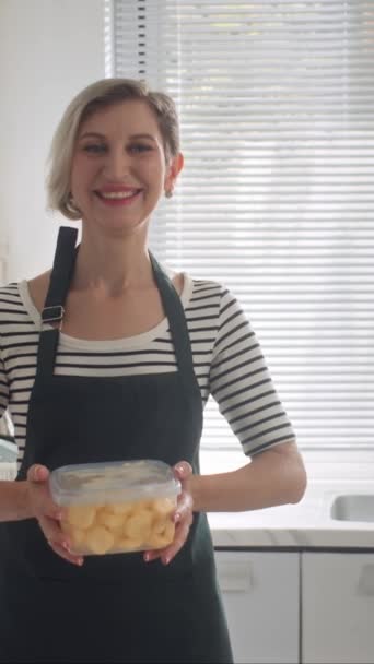 Retrato vertical de alegre ama de casa caucásica mirando a la cámara con contenedor de masa de galletas en la cocina casera - Metraje, vídeo