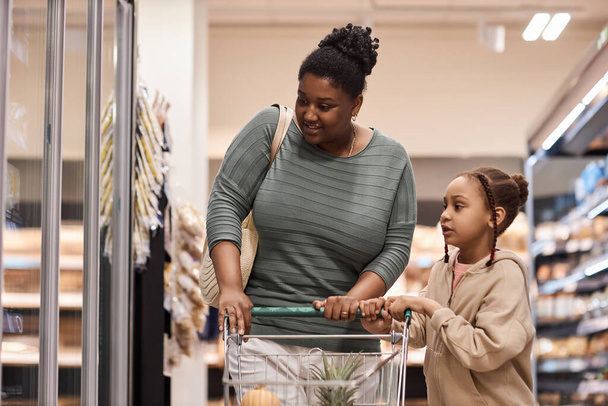 Porträt einer schwarzen Frau, die mit kleinem Mädchen im Supermarkt Lebensmittel einkauft und einen Einkaufswagen schiebt - Foto, Bild