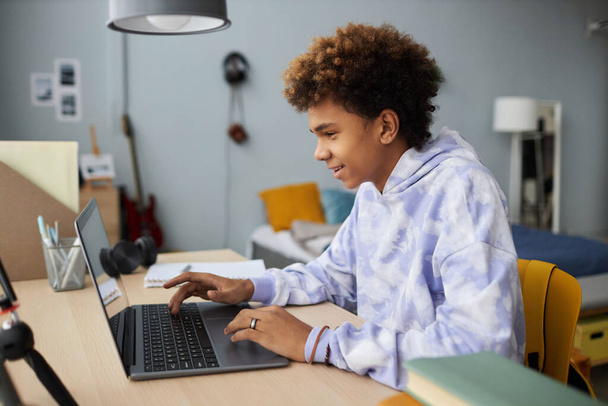 Kapüşonlu genç çocuk bilgisayarda klavyeyle yazı yazıyor ve online ders sırasında öğretmenlerin açıklamalarını dinlerken ekrana bakıyor. - Fotoğraf, Görsel