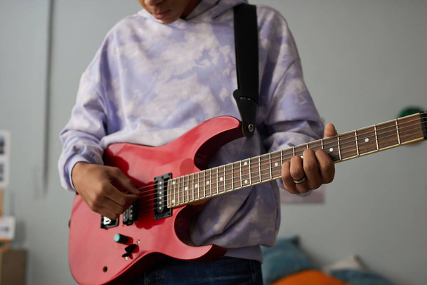 Primer plano del adolescente con sudadera sosteniendo la guitarra eléctrica mientras está parado frente a la cámara y aprendiendo a tocar el instrumento musical de cuerda - Foto, imagen