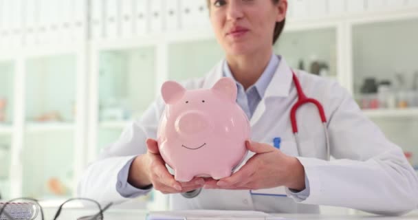 As mãos do médico estão a segurar o porquinho rosa. Doar dinheiro para o conceito de medicina médica - Filmagem, Vídeo