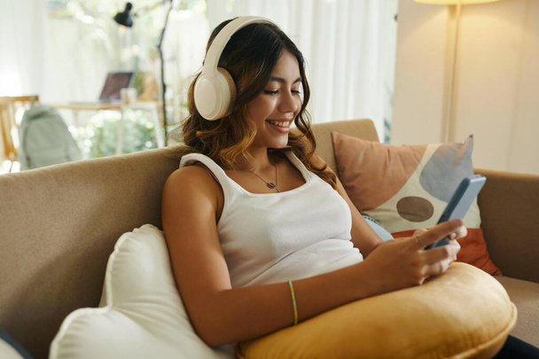 Adolescente souriante dans un casque jouant au jeu sur smartphone lorsqu'elle se repose sur un canapé à la maison - Photo, image