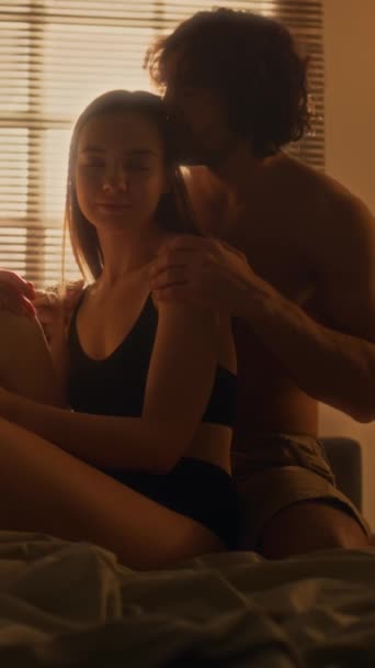 Tiro vertical de joven hombre cariñoso acariciando a su novia en la cama en un acogedor dormitorio con luz cálida - Metraje, vídeo