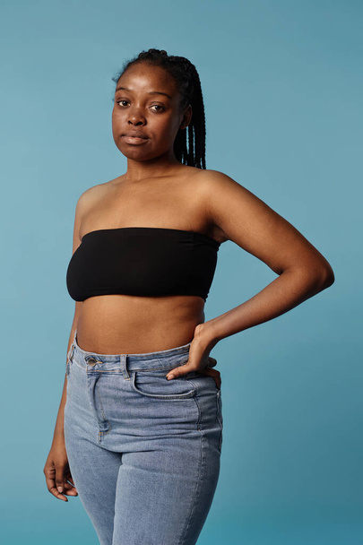 Retrato vertical medio largo de una joven afroamericana con top bandeau y jeans posando para la cámara en el estudio - Foto, imagen