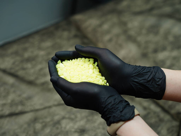 Granulaat goudwas voor ontharing. Depilatiemeester in zwarte beschermende handschoenen houdt korrelige gele ontharingscrème vast - Foto, afbeelding