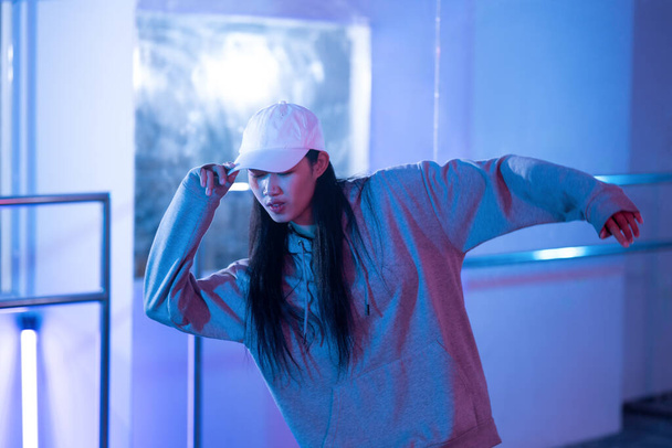 Młoda kobieta w kapturze ćwiczy taniec w studio ze światłami neonowymi. - Zdjęcie, obraz