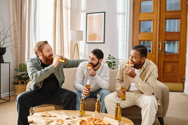 Drie knappe, vrolijke mannen van verschillende rassen zitten op een bank, eten pizza en drinken bier, lachen en genieten. - Foto, afbeelding