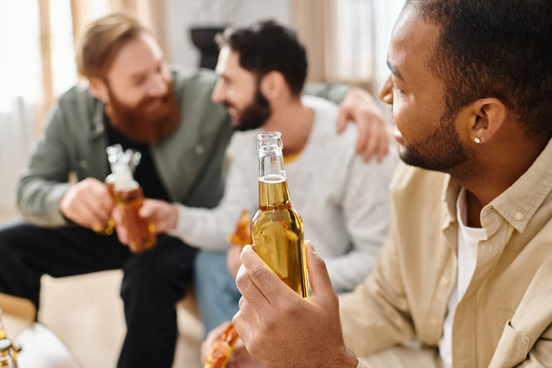 Τρεις ευδιάθετοι, διαφυλετικοί άντρες με καθημερινά ρούχα κάθονται γύρω από ένα τραπέζι, δένονται πίνοντας μπύρες και περνάνε καλά μαζί.. - Φωτογραφία, εικόνα