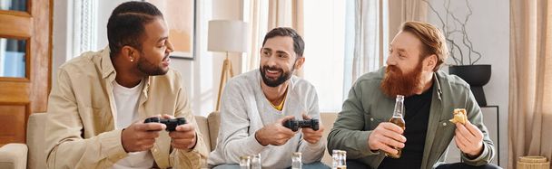 Three cheerful, interracial men sit around a table holding remotes, sharing laughs and camaraderie in a casual setting. - Photo, Image