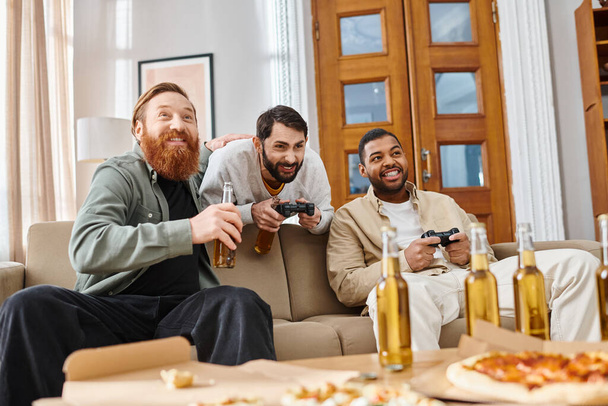 Τρεις όμορφοι άντρες διαφορετικών φυλών μοιράζονται χαμόγελα και συζητήσεις γύρω από ένα τραπέζι γεμάτο πίτσα και μπύρα, απολαμβάνοντας ένα χαλαρό βράδυ φιλίας στο σπίτι.. - Φωτογραφία, εικόνα