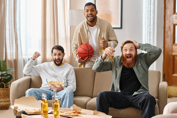 Trzech wesołych, przystojnych mężczyzn różnych ras cieszących się swoim towarzystwem na przytulnej kanapie w domu. - Zdjęcie, obraz
