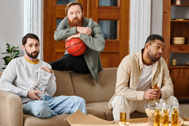 Tres hombres interracial y guapos sentados en un sofá, disfrutando de las bebidas y viendo baloncesto juntos en atuendo casual, mostrando amistad. - Foto, imagen