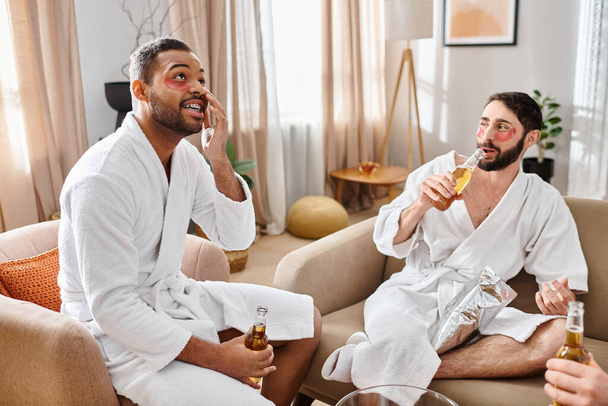 Three diverse, cheerful men in bathrobes enjoying a great time sitting atop a plush couch, sharing laughs and stories. - Photo, Image