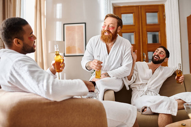 Trois hommes divers et joyeux en peignoirs riant et bavardant dans un cadre confortable de salon. - Photo, image