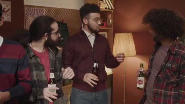 Plan moyen du groupe de jeunes amis masculins du Moyen-Orient debout avec des bouteilles de bière en verre et avoir une conversation pendant la fête de la bière domestique dans un appartement de style rétro avec une lumière jaune chaud confortable - Séquence, vidéo