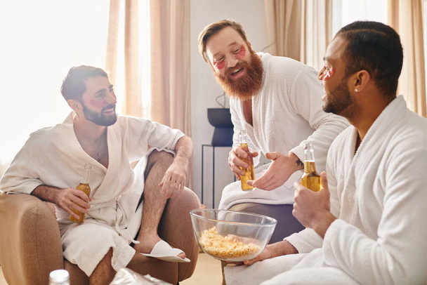 Três homens diversos e alegres em roupões de banho sentados em cima de um sofá, desfrutando uns dos outros da companhia. - Foto, Imagem