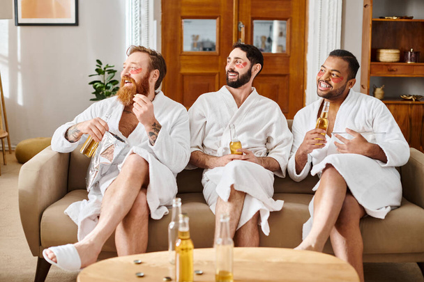 Tre uomini diversi e allegri che indossano accappatoi si stanno divertendo insieme mentre sono seduti su un divano. - Foto, immagini