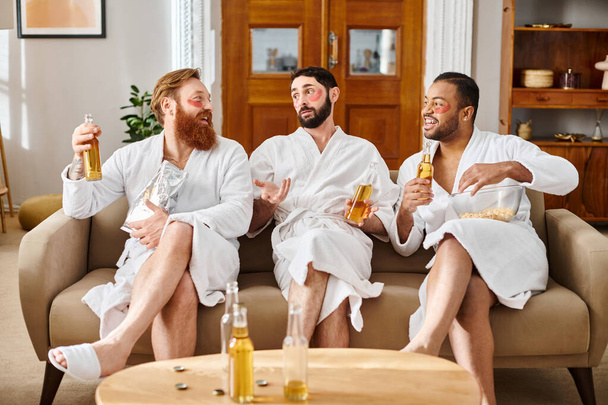 Tres hombres diversos y alegres en albornoces, disfrutando de la compañía de los demás mientras se sientan en la parte superior de un sofá. - Foto, Imagen