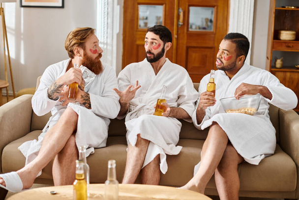 Kolme monipuolista iloinen miesten kylpytakit nauttivat toistensa seurasta istuessaan päälle sohvalla. - Valokuva, kuva