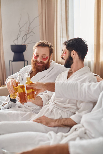 Three diverse and cheerful men in bathrobes enjoy each others company as they sit on top of a sofa, exuding a sense of camaraderie. - Foto, immagini