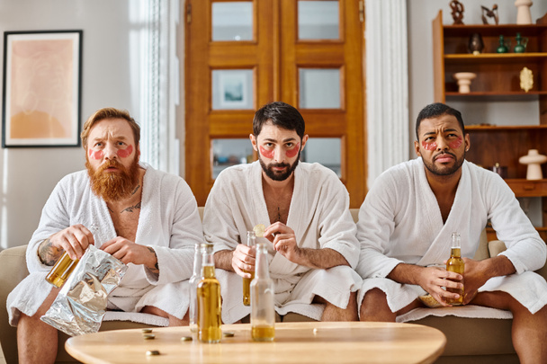 Три задумчивых, разнообразных мужчины в халатах сидят на диване, деля хорошее время и товарищество. - Фото, изображение