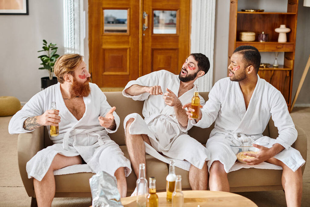 Tres hombres diversos en albornoces se sientan en el sofá, riendo y disfrutando de la compañía de los demás en un momento alegre. - Foto, imagen