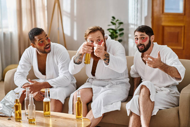 Three cheerful, diverse men in bathrobes sit on top of a couch, sharing laughter and camaraderie. - Photo, Image