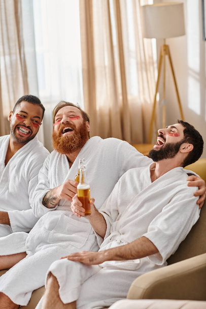 Três homens diversos e alegres em roupões de banho se divertindo sentados juntos em um sofá luxuoso. - Foto, Imagem