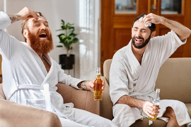 Happy men in bathrobes laugh and chat while perched on top of a couch in a joyful moment of friendship. - Photo, Image