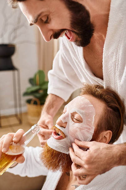 Twee vrolijke mannen in badjassen genieten van een hechtend moment als een zachtjes masker op een ander mans gezicht. - Foto, afbeelding