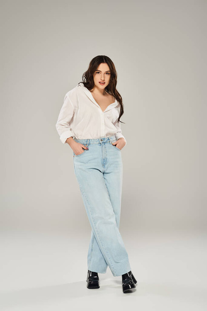 Μια όμορφη γυναίκα συν μέγεθος χτυπά μια στάση σε ένα λευκό πουκάμισο και μπλε τζιν σε ένα γκρι φόντο. - Φωτογραφία, εικόνα