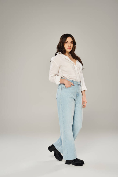 Стильный плюс размер женщина позирует уверенно в белой рубашке и синих джинсах на сером фоне. - Фото, изображение
