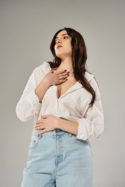 Een prachtige plus size vrouw slaat een pose in een trendy wit shirt en blauwe jeans tegen een neutrale grijze achtergrond. - Foto, afbeelding